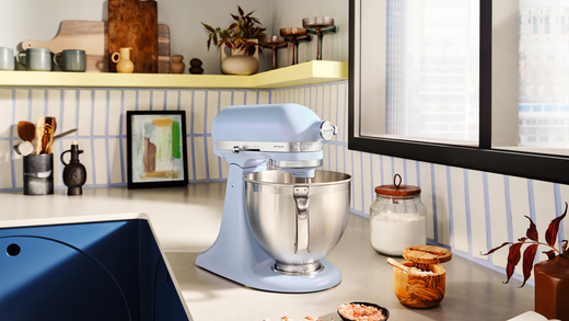 close up of a stand mixer in iridescent blue and lilac colour, photographed on a kitchen worktop surrounded by food supplies and small decorative items