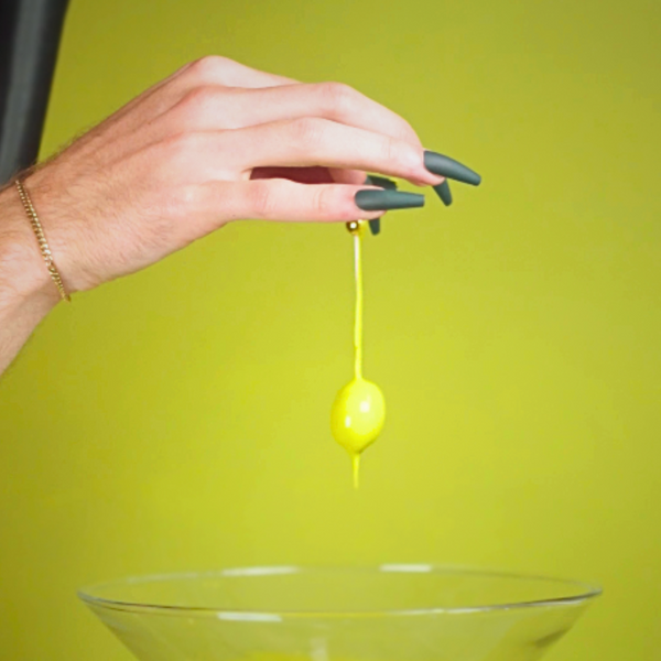 photo of olive green wall with a dark velvet green curtain showing on the left, with a pale skin hand coming out of the corner with dark green long nails, holding an olive that's dripping green paint onto a cocktail glass
