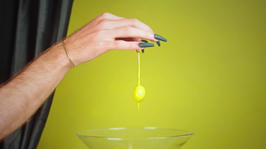 photo of olive green wall with a dark velvet green curtain showing on the left, with a pale skin hand coming out of the corner with dark green long nails, holding an olive that's dripping green paint onto a cocktail glass