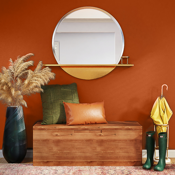 photo of a large rusty orange terracotta wall, with a wooden bench placed on top of it, decorated with green and orange cushions, pampas grass on the left and rain boots and umbrella to the right, with a round mirror above it