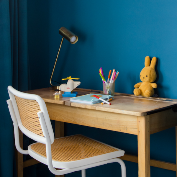 photo of a children's bedroom cover with a vintage wooden desk, white and rattan chair as well as a plush bunny toy, crayons and a table lamp placed on top of it, surrounded by a dark teal colour on the walls and a peachy pink colour on the ceiling