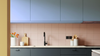 Modern kitchen interior with blue-gray cabinets, a blush pink tiled backsplash, a white countertop, and a black faucet. Decorative white containers, a wooden cutting board, and orange flowers are visible on the counter.