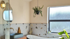 A modern bathroom interior with a freestanding white bathtub, a rectangular blue vessel sink, a wall-mounted mirror, a hanging green plant, and a large frosted window. The wall features a terrazzo-style tile pattern in beige, gray, and brown tones.