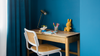 A wooden writing desk with a white woven-back chair in a styled room corner, set against a deep blue wall with a matching curtain. The desk is styled with a black desk lamp, a cup of colored pencils, and a yellow plush bunny.