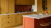 A modern kitchen interior with mustard yellow cabinets, a terracotta tiled backsplash, white countertops, and a central island in coral orange with wooden stools.