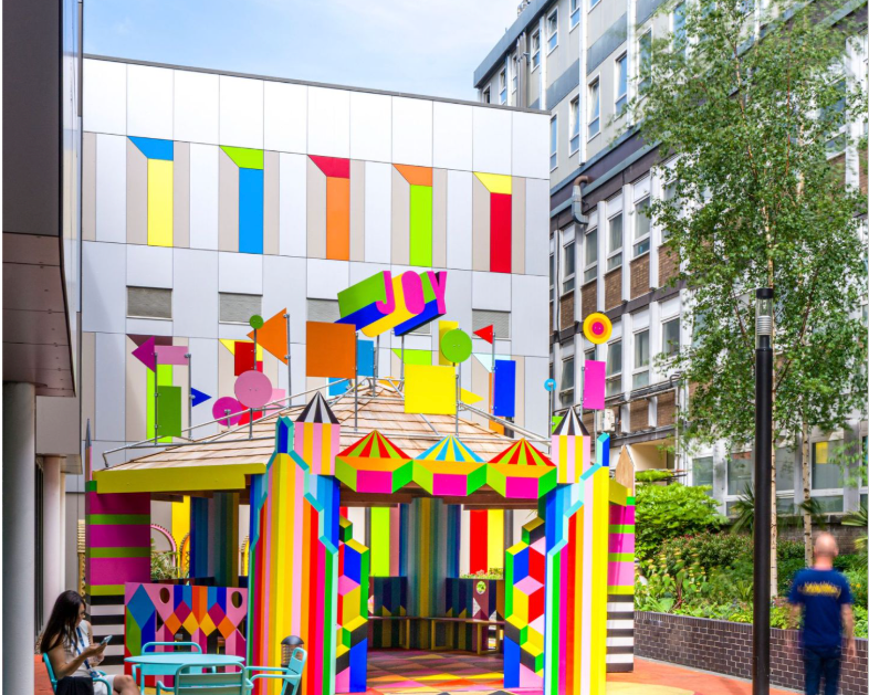 A vibrant, colorful urban courtyard featuring a whimsical carousel-like pavilion with bold geometric patterns, colorful tiled flooring, and modern buildings in the background. A few people are visible in the scene.