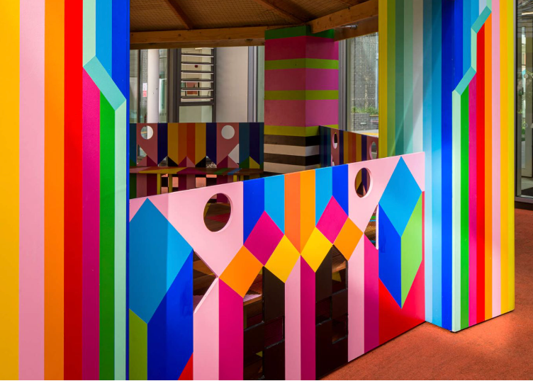A colorful architectural structure with bold geometric wall panels and a bright yellow roof overhang, set in front of a modern glass building.