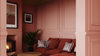 A cozy living room corner with dusty rose paneled walls, a rust-red sofa with striped pillows, a potted green plant, and a fireplace on the left.