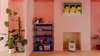 A pink room interior with a blue shelving unit holding small items, potted plants, a framed wall art print, and a small toy house on the floor.