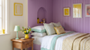 A cozy bedroom scene with a bed dressed in white and pale green bedding, set against a lavender wood-paneled wall. A wicker nightstand with a mint green lamp and a vase of flowers sits beside the bed, and framed yellow artwork hangs on the wall.