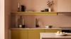A modern kitchen interior with mustard yellow cabinets and floating shelves against a warm peach-pink wall. A black faucet, bowls, a candle holder, and a vase with dried stems are arranged on the shelves and counter.