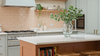 A modern kitchen interior with pale green cabinets, a peach subway tile backsplash, a white island countertop, and a clear glass vase holding green eucalyptus leaves on top.