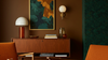 A styled interior scene with a mid-century wooden sideboard, white dome table lamp, wall sconce, framed abstract artwork, books, and orange upholstered chairs against a dark brown wall.
